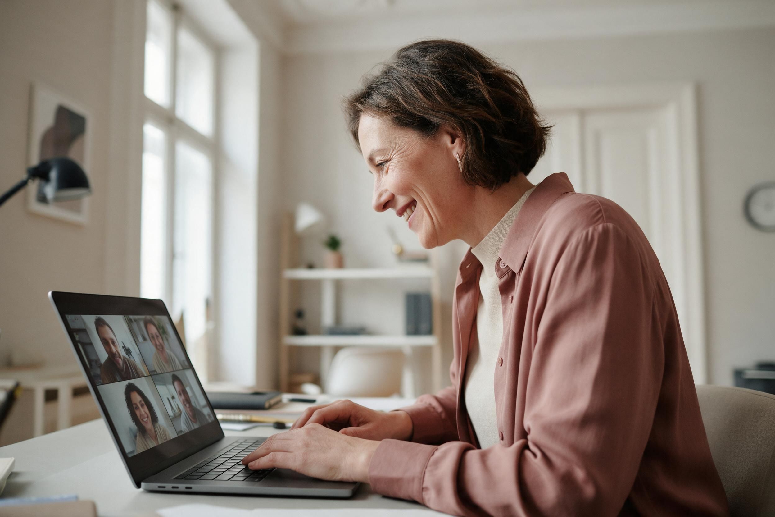Eine lächelnde Frau sitzt am Schreibtisch und führt am Laptop ein Videogespräch mit einer Kleingruppe.
